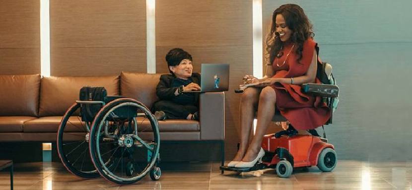Resim: Kahverengi deri 3'lü koltuğun ortasında boş tekerlekli sandalye var. Koltuğun sağ tarafına oturmuş ortopedik engeli olan kadın koltuk koluna koyduğu dizüstü bilgisayarında çalışıyor. Koltuğun sağında da tekerlekli sandalyede oturan kadın ile hem sohbet ediyorlar hem de çalışıyorlar.
