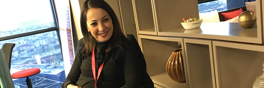Betimleme: Resimde modern bir ofis ortamı görülüyor. Ön planda siyah kıyafetli, kırmızı yaka kartı takan bir kişi oturuyor. Arkadaki raflarda dekoratif objeler (altın vazo, bitkili kase vb.) bulunuyor. Büyük pencereden şehir manzarası ve iç mekânda kırmızı bir koltuk dikkat çekiyor.