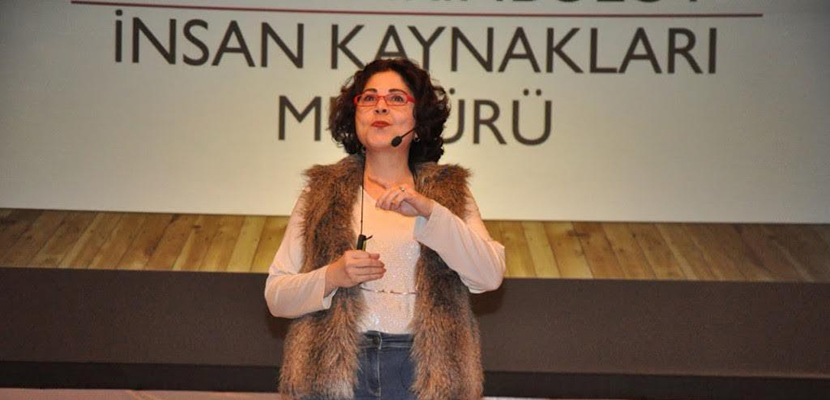 Görselde, sahnede sunum yapan Elif Yıldırımbulut görülüyor. Kırmızı gözlükleri, beyaz gömleği ve kahverengi kürk yeleğiyle dikkat çekiyor. Kulaklık mikrofonu takılı ve arka planda “İnsan Kaynakları Müdürü” yazısı yer alıyor.