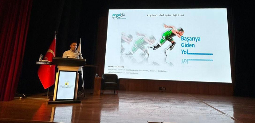 Betimleme: Sahnenin ortasında bir kürsüde Mehmet Kızıltaş ayakta duruyor. Arka planda büyük bir projeksiyon perdesi var; üzerinde protez bacaklı bir atletin koşarken çekilmiş fotoğrafı yer alıyor. Slaytta “Kişisel Gelişim Eğitimi” ve “Başarıya Giden Yol” gibi ifadeler bulunuyor. Sahnede Türk bayrağı ve üniversitenin bayrağı da var. Mekân geniş, aydınlık ve resmi bir konferans salonu havasında. Mehmet Kızıltaş, Engelsizkariyer.com’un kurucusu olarak katılımcılara hitap ediyor.