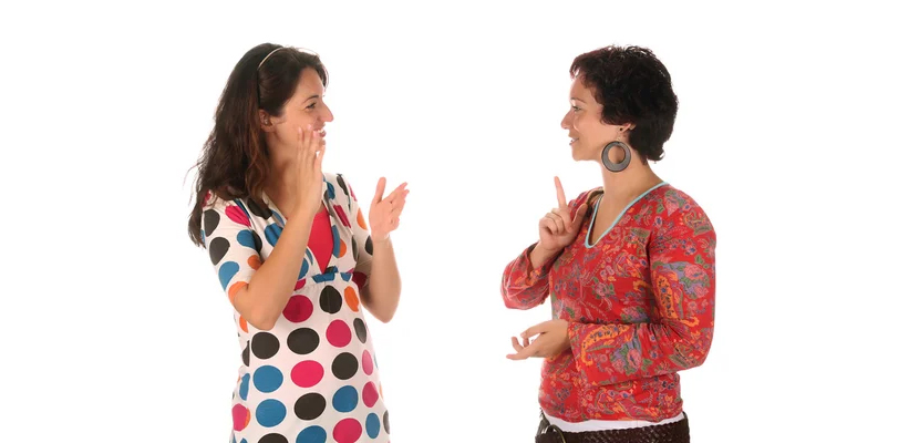 Betimleme: İki kişi düz beyaz arka plan önünde işaret diliyle iletişim kuruyor; yüz ifadeleri ve el hareketleri belirgin.