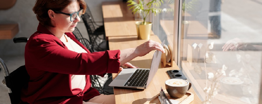 Betimleme: Kırmızı ceket giymiş cam kenarın masada çalışmak için laptop bilgisayarını açarken sağ profilinden görünen tekerlekli sandalye kullanıcısı kadın fotoğrafı.