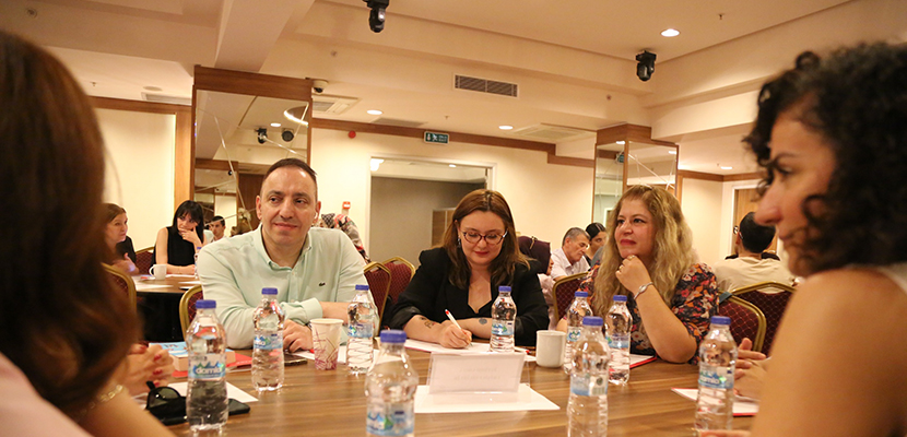 Yuvarlak masada toplanmış küçük bir grup, notlar ve su şişeleri arasında ciddi bir toplantı yapıyor; ortam profesyonel ve odaklı.