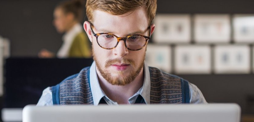 Modern bir ofiste, masasında dizüstü bilgisayarla çalışan bir kişi var. Gözlüklü, gömlek ve süveter yelek giymiş. Masada birkaç kâğıt ve kalem duruyor. Arka planda iki kişi ayakta sohbet ediyor ve duvarda düzenli şekilde asılmış çerçeveler bulunuyor. Ortam aydınlık, düzenli ve profesyonel bir çalışma alanı hissi veriyor.