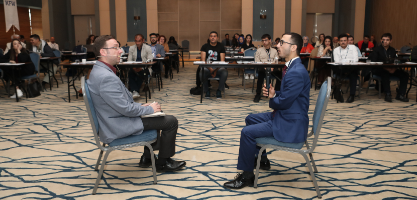 Resim: Eğitim salonunda sıralarda oturan engeli olan katılımcılar sahnede İK'cı ve engeli olan adayın mülakat simülasyonunu takip ediyor.