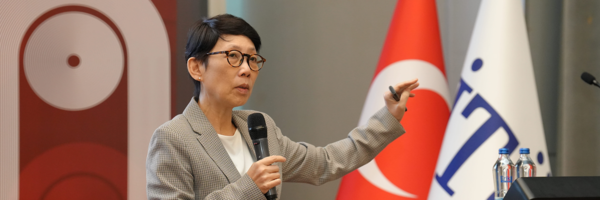 Resim Betimleme: Bir konferans salonunda, kürsü önünde konuşma yapan bir erkek görülüyor. Üzerinde gri tonlarda kareli bir blazer ceket ve gözlük bulunuyor. Elinde mikrofon tutarak konuşuyor ve diğer eliyle vurgu yapar şekilde jest yapıyor. Arka planda kırmızı zemin üzerinde beyaz daireli bir bayrak ile yanında beyaz zemin üzerinde mavi yazılı bir bayrak yer alıyor. Kürsünün üzerinde Erikli marka iki su şişesi bulunuyor. Genel atmosfer, akademik bir sunum veya resmi bir eğitim etkinliği izlenimi veriyor.