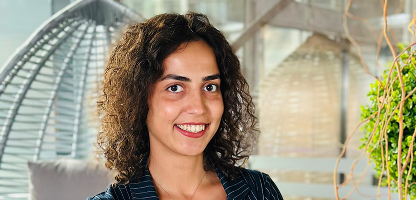Resimde, koyu renk çizgili bir takım elbise ve beyaz bir üst giyen, kendinden emin duruşlu Sena Fıstıkçık görülüyor. Kolları bağlı, modern ve şık bir iç mekânda ayakta duruyor. Arka planda cam duvarlar, bitkiler ve dekoratif bir salıncak koltuk bulunuyor.