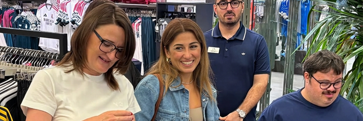 Mağaza içinde, spor giyim ürünlerinin bulunduğu rafların önünde 3 kişi yan yana 1 kişi de arkada duruyor. Solda elinde parlak bir paket tutan Michelle Robertson, yanında kot ceketli İrem Önal ve sağda down sendromlu Berk Can Bulat yer alıyor. Hemen arkalarında lacivert tişörtüyle mağaza yöneticisi bir erkek bulunuyor. Grup, mağaza içindeki samimi ve profesyonel atmosferde kameraya dönük şekilde poz veriyor.